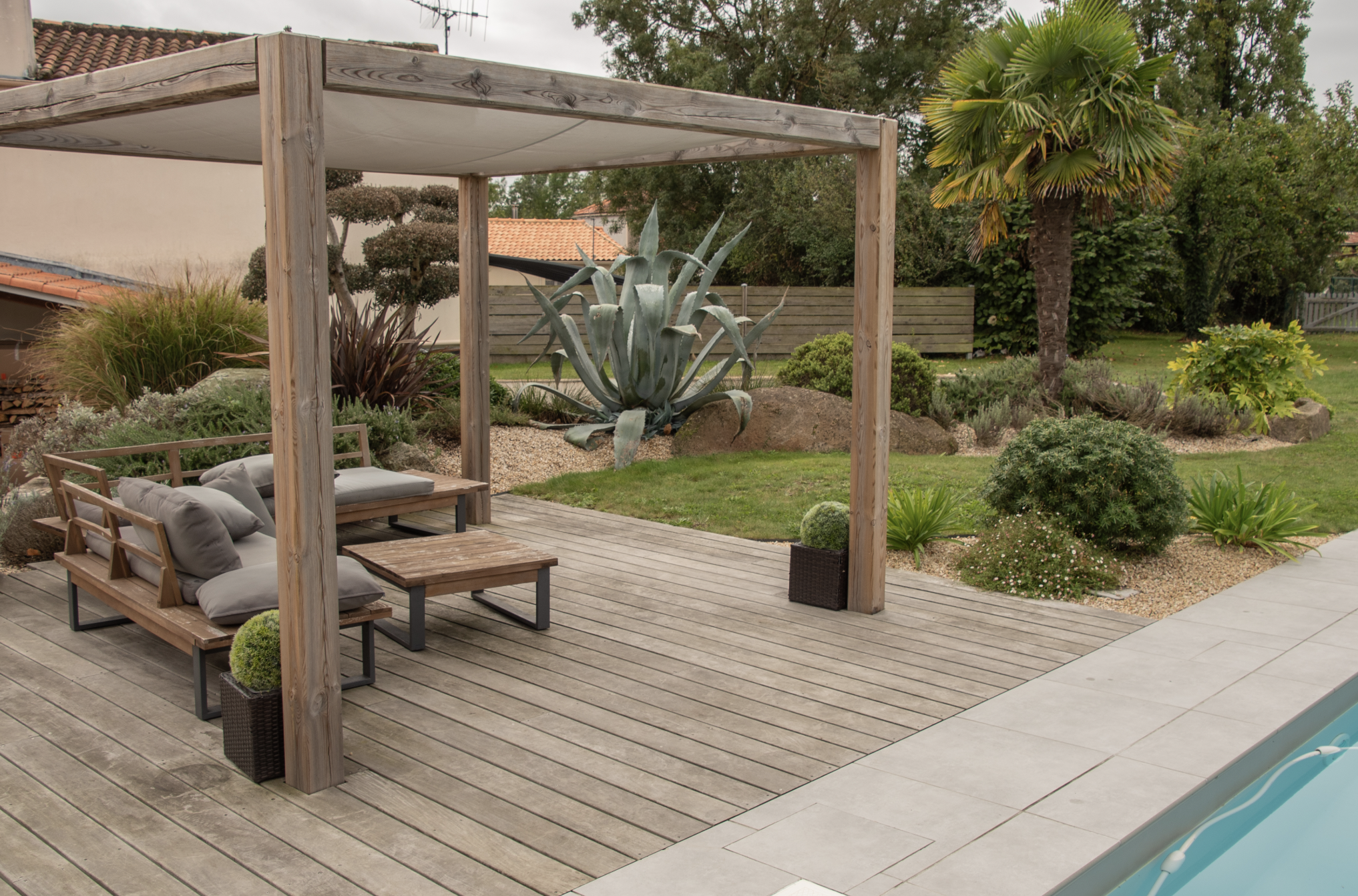 Terrasse en bois exotique avec pergola et salon de jardin, aménagée au bord d’une piscine dans un jardin paysager moderne à Clisson.