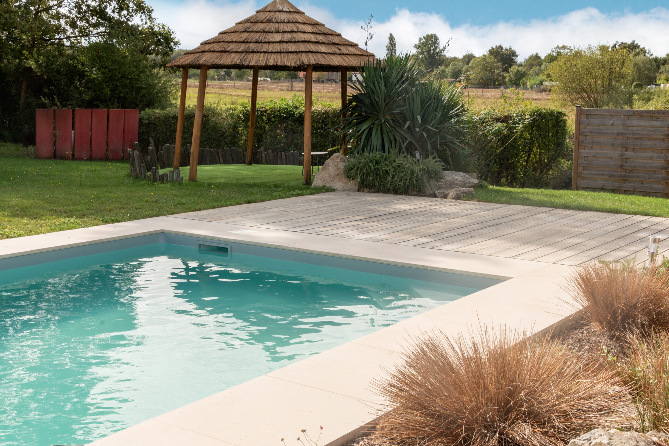 Aménagement extérieur avec piscine et terrasse en bois, margelles en pierre et paillote en bois, intégré dans un jardin paysager naturel par un paysagiste en Vendée.
