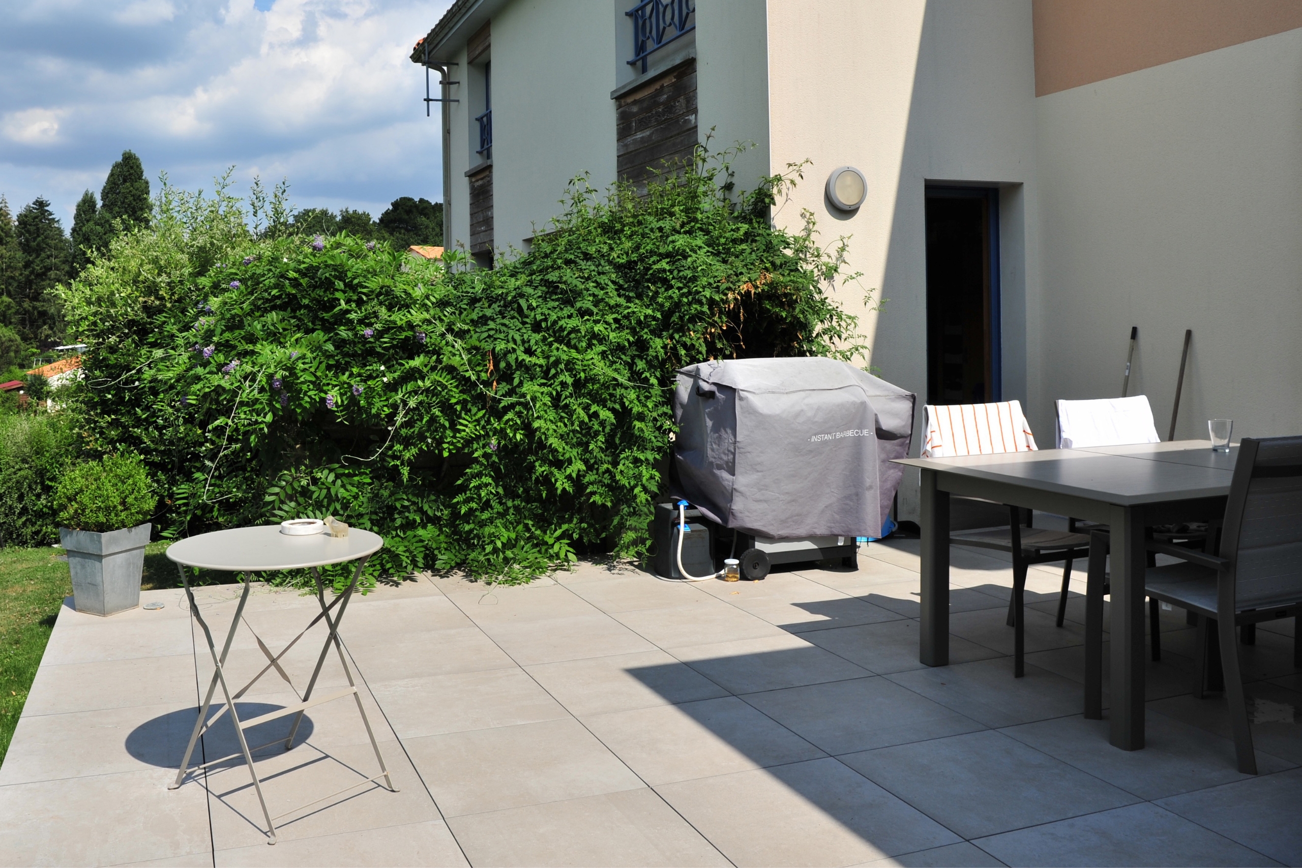 Terrasse contemporaine réalisée en dalles de grès cérame grand format de couleur beige. L'espace comprend un salon de jardin moderne, un barbecue protégé et une haie de verdure dense bordant la maison.