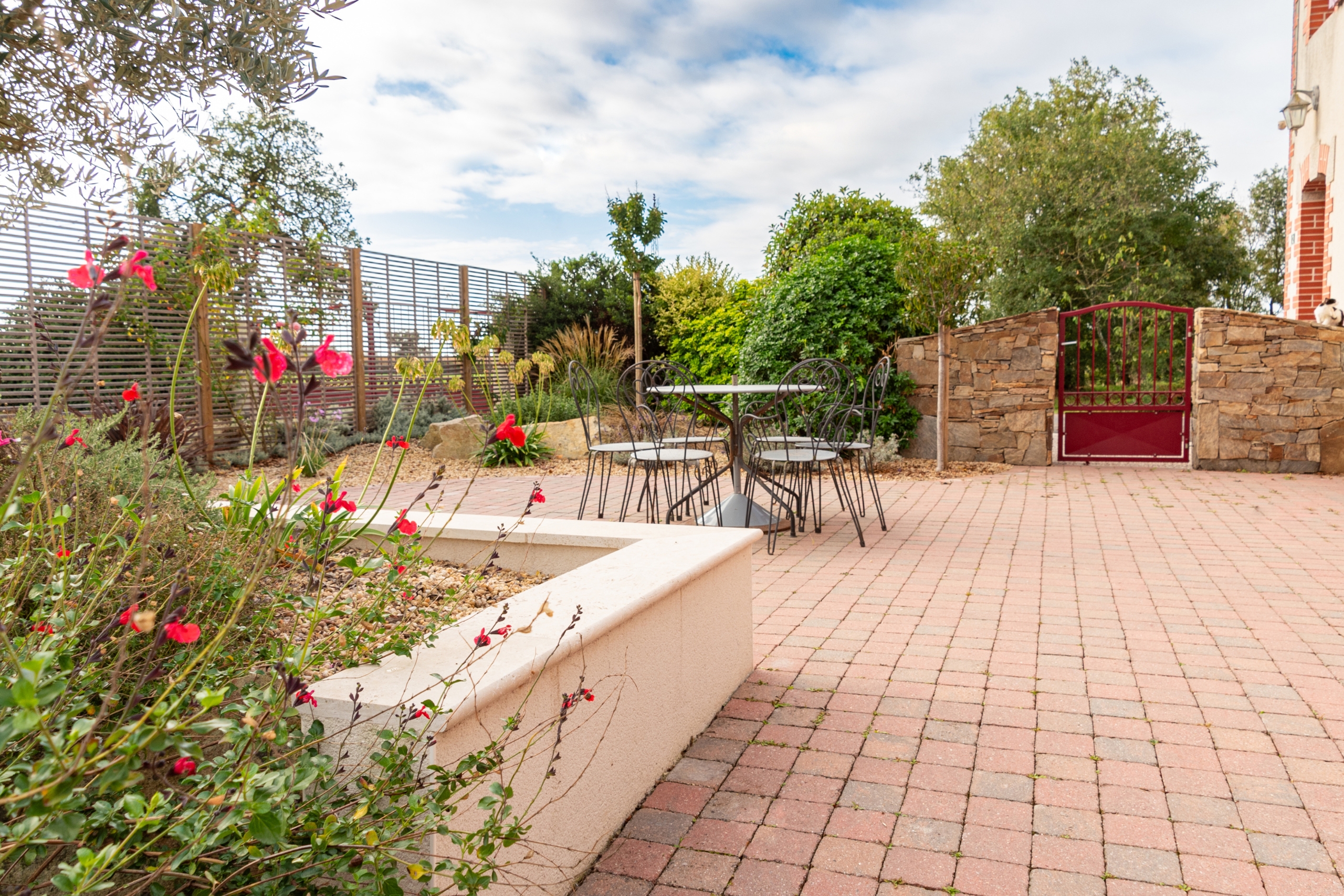 Aménagement d'une terrasse authentique en pavés roses avec un salon de jardin en fer forgé. L'espace est délimité par des murets en pierre naturelle, un portillon rouge et des plantations méditerranéennes.